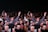 A large group of people gathered in a celebratory atmosphere, many smiling and raising their hands. The central figure is hoisted above the crowd, suggesting a lively, energetic event.