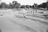 A bustling skatepark in a park setting, with numerous people engaged in skateboarding and other activities. The area is filled with ramps, rails, and concrete platforms, surrounded by lush trees and grassy areas. A lamppost stands in the center, and people are both actively skating and watching from the sidelines.
