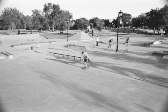 A bustling skatepark in a park setting, with numerous people engaged in skateboarding and other activities. The area is filled with ramps, rails, and concrete platforms, surrounded by lush trees and grassy areas. A lamppost stands in the center, and people are both actively skating and watching from the sidelines.