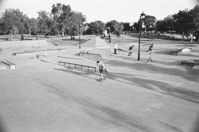 A bustling skatepark in a park setting, with numerous people engaged in skateboarding and other activities. The area is filled with ramps, rails, and concrete platforms, surrounded by lush trees and grassy areas. A lamppost stands in the center, and people are both actively skating and watching from the sidelines.