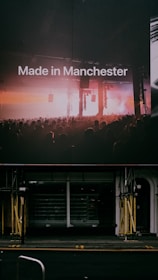 A large billboard displays the text 'Made in Manchester' in front of a concert scene with a crowd of people. Bright lights illuminate the stage area, casting a warm glow. Below the billboard, there is a dark, vacant storefront with scaffolding on either side.