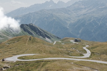 The image features a winding road traversing through a mountainous landscape with green grass and rocky terrain. Clouds can be seen hanging low on the left side while high, rugged mountain peaks are in the background. A small building is situated along the road in the distance.