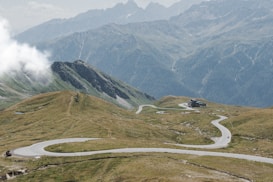 The image features a winding road traversing through a mountainous landscape with green grass and rocky terrain. Clouds can be seen hanging low on the left side while high, rugged mountain peaks are in the background. A small building is situated along the road in the distance.