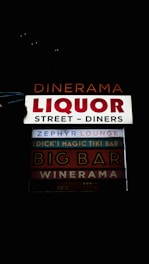 A series of illuminated signs advertising various establishments, including bars and diners. The signs are arranged vertically and feature names like 'Dinerama,' 'Liquor Street - Diners,' and 'Zephyr Lounge,' among others. The lettering is bold and colorful against a dark background, implying a nighttime setting.