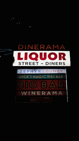 A series of illuminated signs advertising various establishments, including bars and diners. The signs are arranged vertically and feature names like 'Dinerama,' 'Liquor Street - Diners,' and 'Zephyr Lounge,' among others. The lettering is bold and colorful against a dark background, implying a nighttime setting.