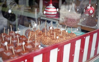 A display of caramel apples with wooden sticks is arranged neatly behind a glass window. Some apples are coated with nuts. The display features a retro-style 'Yes, we are open' sign, and two Christmas-themed ornaments hang from the top. The environment suggests a festive, holiday atmosphere.