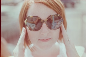 A person with short hair wearing oversized, stylish sunglasses, holding the frames gently with both hands. The background is softly blurred, giving a dreamy, vintage feel to the image.