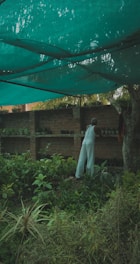 A person dressed in white is tending to plants in a lush, green garden, surrounded by a variety of foliage and potted plants. The area is shaded by a large green net hanging above, providing a tranquil and peaceful atmosphere.
