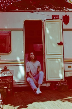 A woman sits casually in the open doorway of a vintage camper van, sticking out her tongue playfully. She is dressed in casual attire with light-colored pants and sneakers, holding a drink in one hand. The setting is outdoors with a wooden deck and a few potted plants nearby. The lighting has a reddish tint, giving the image a warm, nostalgic feel.