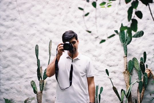 A man wearing a white polo shirt is holding a camera and taking a photograph. He is standing in front of a textured white wall, surrounded by several tall cactus plants. Some leaves can be seen hanging from above.
