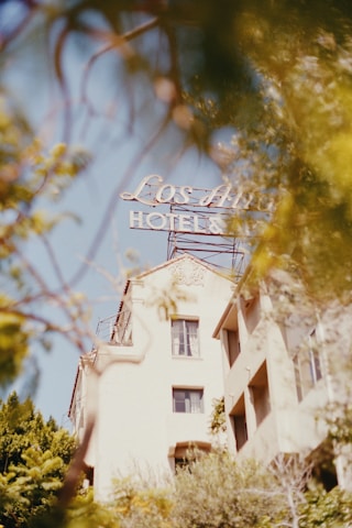 A vintage-style photograph of a hotel building framed by foliage, featuring a sign that reads 'Los Angeles Hotel' in classic lettering. The structure has a nostalgic architectural style, with soft details and pastel colors, partially obscured by branches and leaves, adding a dreamy effect.