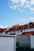 A modern building complex with multiple pitched roofs in a vibrant orange color. The structure has several dormer windows, and the facade contrasts with the bright blue sky above. A separate garage or storage unit marked with the number '34' is visible in the foreground, with a barrier wall to its side.