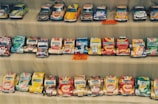 Rows of handmade toy cars crafted from recycled tin cans are neatly displayed. Each car features colorful and recognizable branding from various beverage companies. The setup appears to be part of a market or street vendor display with small price tags visible.