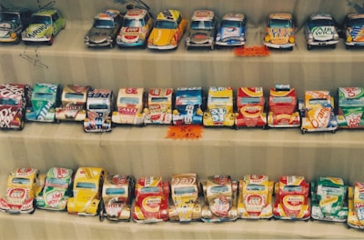 Rows of handmade toy cars crafted from recycled tin cans are neatly displayed. Each car features colorful and recognizable branding from various beverage companies. The setup appears to be part of a market or street vendor display with small price tags visible.