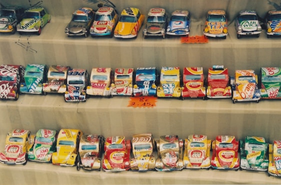 Rows of handmade toy cars crafted from recycled tin cans are neatly displayed. Each car features colorful and recognizable branding from various beverage companies. The setup appears to be part of a market or street vendor display with small price tags visible.