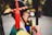 A person in traditional Korean attire is facing sideways. The hat features a red and yellow braided band and tassel. The clothing is colorful, comprising green, yellow, and blue, suggesting a ceremonial or traditional event. The background is blurred, indicating an outdoor setting.
