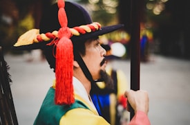 A person in traditional Korean attire is facing sideways. The hat features a red and yellow braided band and tassel. The clothing is colorful, comprising green, yellow, and blue, suggesting a ceremonial or traditional event. The background is blurred, indicating an outdoor setting.