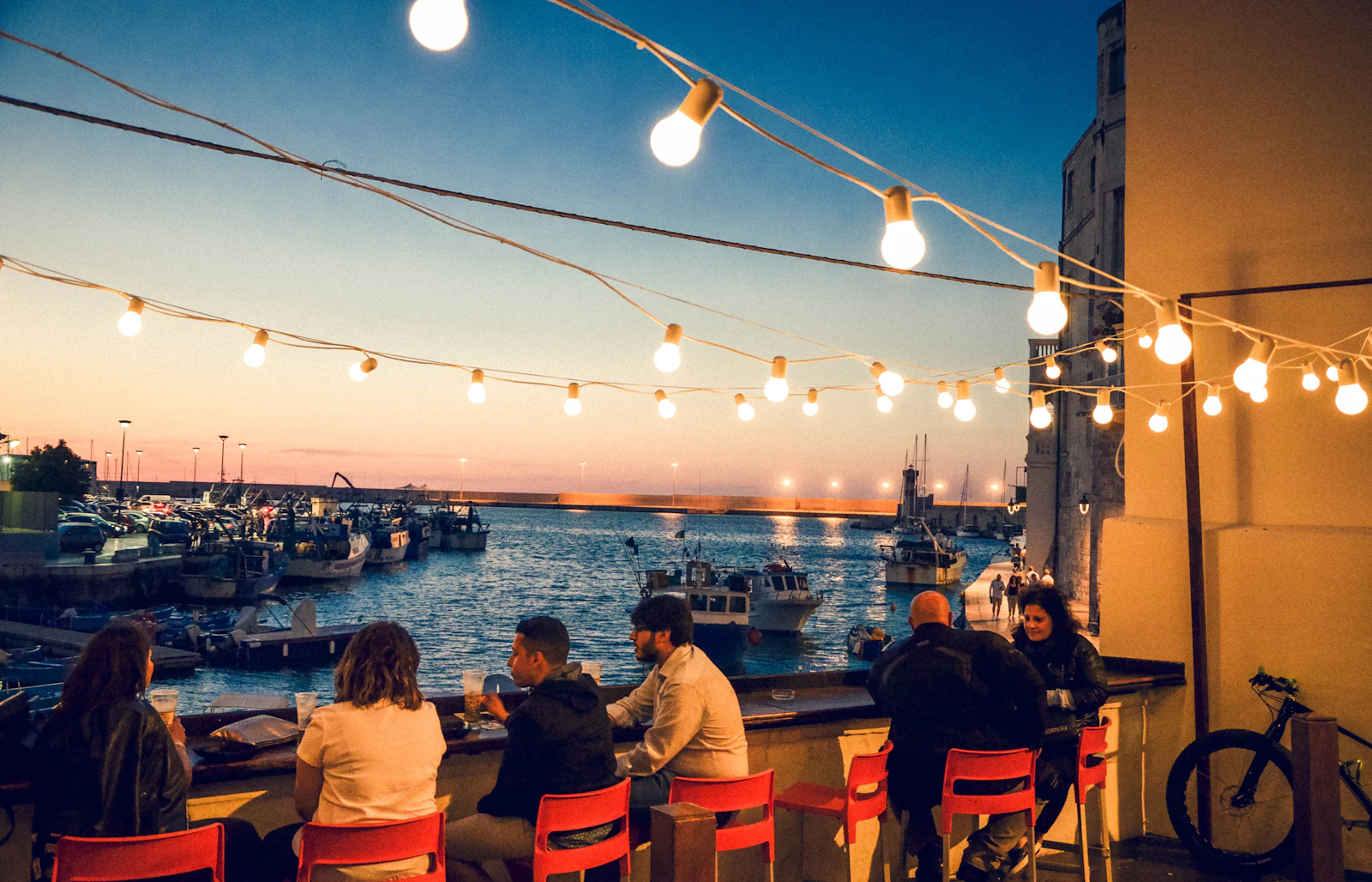 People sitting along a harbour wall under strings of lit bulbs at dusk, fishing boats moored below them.