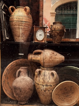 A collection of vintage pottery and ceramics is displayed in a window. The arrangement includes large amphoras with decorative patterns, a round clock with a wooden frame, and various plates all exhibiting earthy tones. The background features a partially visible arched window with a green shutter.