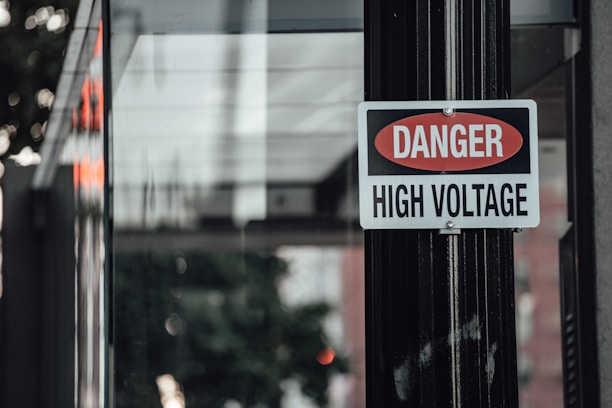 A sign attached to a black pole with the words 'Danger High Voltage' in bold red and black text. The background shows a glass window with reflections and blurry green foliage.