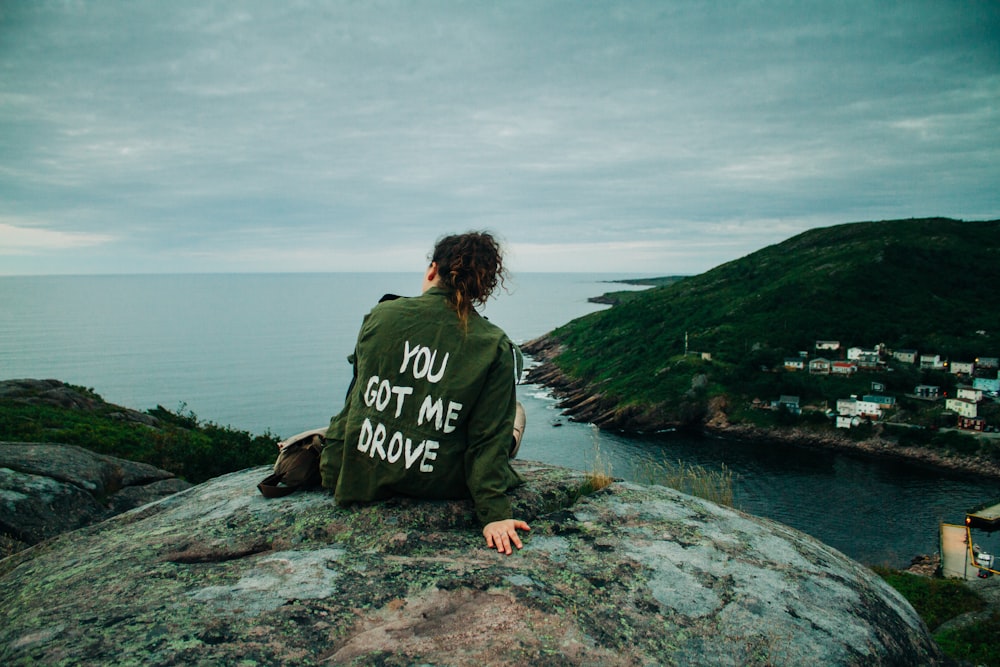 woman in gray and black long sleeve shirt sitting on gray rock near body of water