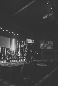A black and white photograph captures a live music performance on a stage with a band. A large camera is set up in the foreground, indicating it might be a recording or live broadcast setup. There are musicians with musical instruments such as a guitar and drums. The background features vertical light bars, and a large screen displaying the stage setup.