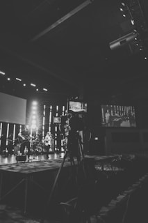 A black and white photograph captures a live music performance on a stage with a band. A large camera is set up in the foreground, indicating it might be a recording or live broadcast setup. There are musicians with musical instruments such as a guitar and drums. The background features vertical light bars, and a large screen displaying the stage setup.