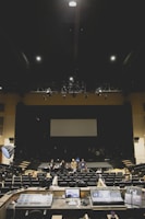 A large, dimly lit auditorium with several people standing on stage and seated in the audience. In the foreground, various audio and lighting control equipment is visible. The room has rows of empty seats and a large screen at the back of the stage.