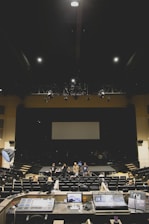 A large, dimly lit auditorium with several people standing on stage and seated in the audience. In the foreground, various audio and lighting control equipment is visible. The room has rows of empty seats and a large screen at the back of the stage.