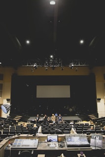 A large, dimly lit auditorium with several people standing on stage and seated in the audience. In the foreground, various audio and lighting control equipment is visible. The room has rows of empty seats and a large screen at the back of the stage.