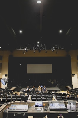 A large, dimly lit auditorium with several people standing on stage and seated in the audience. In the foreground, various audio and lighting control equipment is visible. The room has rows of empty seats and a large screen at the back of the stage.