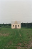 A solitary, small house with a steeply pitched roof and cream-colored walls, situated in a vast open field of green grass. The house is centered in front of a backdrop of dense trees, creating a contrast between the structured building and the natural surroundings.