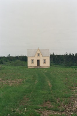 A solitary, small house with a steeply pitched roof and cream-colored walls, situated in a vast open field of green grass. The house is centered in front of a backdrop of dense trees, creating a contrast between the structured building and the natural surroundings.