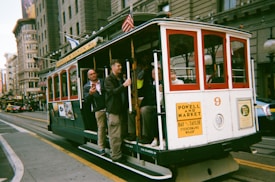 A classic San Francisco cable car travels along a city street with passengers on board. The cable car features red and white colors and is marked with signage indicating routes to places like Powell and Market. Many people are standing on the side of the cable car, holding onto poles while others are seated inside. Urban buildings and city activity are visible in the background.