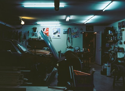 A cluttered garage with a car parked inside, its hood open. Various tools and equipment are scattered around, including a bicycle wheel, workbench, and several posters adorning the walls. The lighting is dim and illuminated by fluorescent ceiling lights, creating a moody atmosphere.