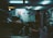 A cluttered garage with a car parked inside, its hood open. Various tools and equipment are scattered around, including a bicycle wheel, workbench, and several posters adorning the walls. The lighting is dim and illuminated by fluorescent ceiling lights, creating a moody atmosphere.