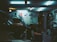A cluttered garage with a car parked inside, its hood open. Various tools and equipment are scattered around, including a bicycle wheel, workbench, and several posters adorning the walls. The lighting is dim and illuminated by fluorescent ceiling lights, creating a moody atmosphere.