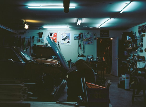 A cluttered garage with a car parked inside, its hood open. Various tools and equipment are scattered around, including a bicycle wheel, workbench, and several posters adorning the walls. The lighting is dim and illuminated by fluorescent ceiling lights, creating a moody atmosphere.