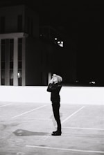 A shadowy figure observing a city street through binoculars at dusk.