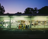 A laundromat is seen under fading evening light, with bright fluorescent lights illuminating the interior. The windows display lettering advertising services like wash, dry, and fold. Outside, newspaper and vending machines are positioned beside the entrance. Trees and a deepening purple sky form the backdrop.