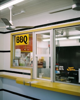 A small takeout window with a yellow sign that reads 'BBQ'. The area is enclosed with glass, showing a counter inside with various small items, such as a cup and papers. There are signs displaying operational hours and payment options. The exterior counter is yellow and there are black stripes on the walls. A ceiling fan is visible inside as well.