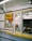 A small takeout window with a yellow sign that reads 'BBQ'. The area is enclosed with glass, showing a counter inside with various small items, such as a cup and papers. There are signs displaying operational hours and payment options. The exterior counter is yellow and there are black stripes on the walls. A ceiling fan is visible inside as well.