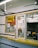 A small takeout window with a yellow sign that reads 'BBQ'. The area is enclosed with glass, showing a counter inside with various small items, such as a cup and papers. There are signs displaying operational hours and payment options. The exterior counter is yellow and there are black stripes on the walls. A ceiling fan is visible inside as well.