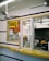 A small takeout window with a yellow sign that reads 'BBQ'. The area is enclosed with glass, showing a counter inside with various small items, such as a cup and papers. There are signs displaying operational hours and payment options. The exterior counter is yellow and there are black stripes on the walls. A ceiling fan is visible inside as well.
