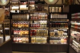 Shelves stocked with various jars, bottles, and packages of sauces are organized in neat rows. The shelves are wooden with a dark finish, and the products include different types of condiments and jams. A person is partially visible on the left side, blending into the display setting.