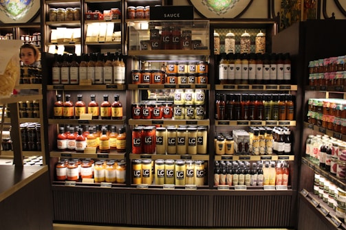 Shelves stocked with various jars, bottles, and packages of sauces are organized in neat rows. The shelves are wooden with a dark finish, and the products include different types of condiments and jams. A person is partially visible on the left side, blending into the display setting.