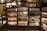Shelves stocked with various jars, bottles, and packages of sauces are organized in neat rows. The shelves are wooden with a dark finish, and the products include different types of condiments and jams. A person is partially visible on the left side, blending into the display setting.