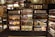 Shelves stocked with various jars, bottles, and packages of sauces are organized in neat rows. The shelves are wooden with a dark finish, and the products include different types of condiments and jams. A person is partially visible on the left side, blending into the display setting.