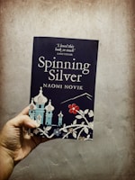 A hand holds a book titled 'Spinning Silver' by Naomi Novik. The cover features stylized illustrations of buildings with onion-shaped domes and floral designs. A quote from Laini Taylor is visible at the top.