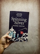 A hand holds a book titled 'Spinning Silver' by Naomi Novik. The cover features stylized illustrations of buildings with onion-shaped domes and floral designs. A quote from Laini Taylor is visible at the top.